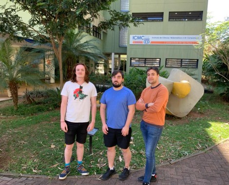 Three white males stand in front of trees and a building.